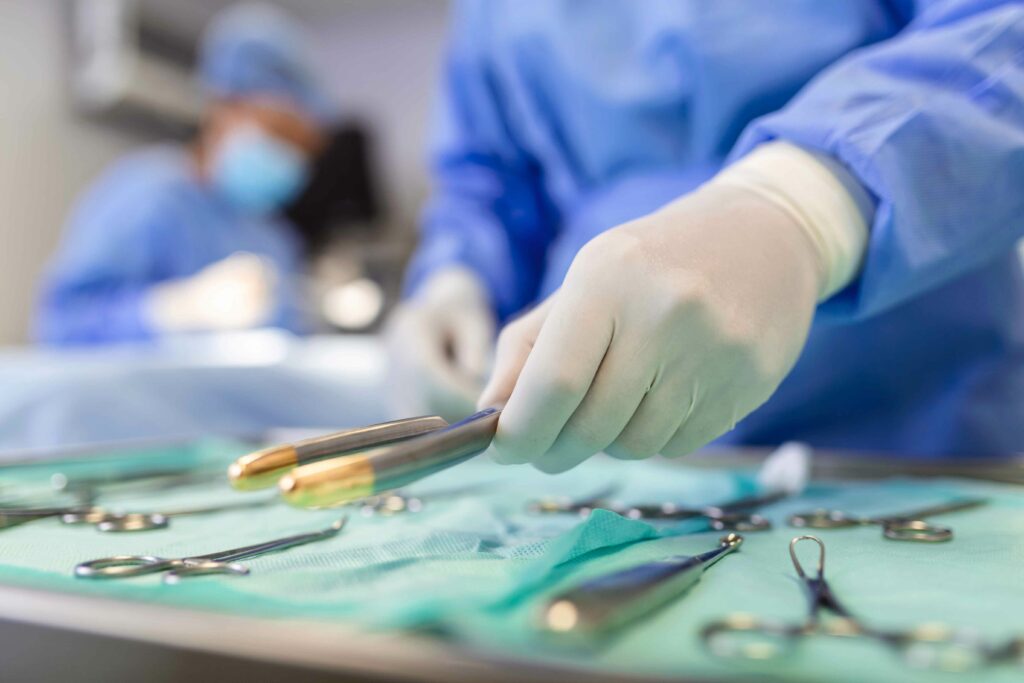 Mão de cirurgião segurando instrumento cirúrgico esterilizado durante cirurgia de quadril, com outros instrumentos em uma bandeja cirúrgica em segundo plano.