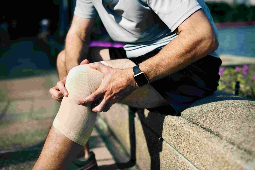 Homem sentado ao ar livre segurando o joelho com uma bandagem elástica, indicando dor ou recuperação de uma lesão esportiva.