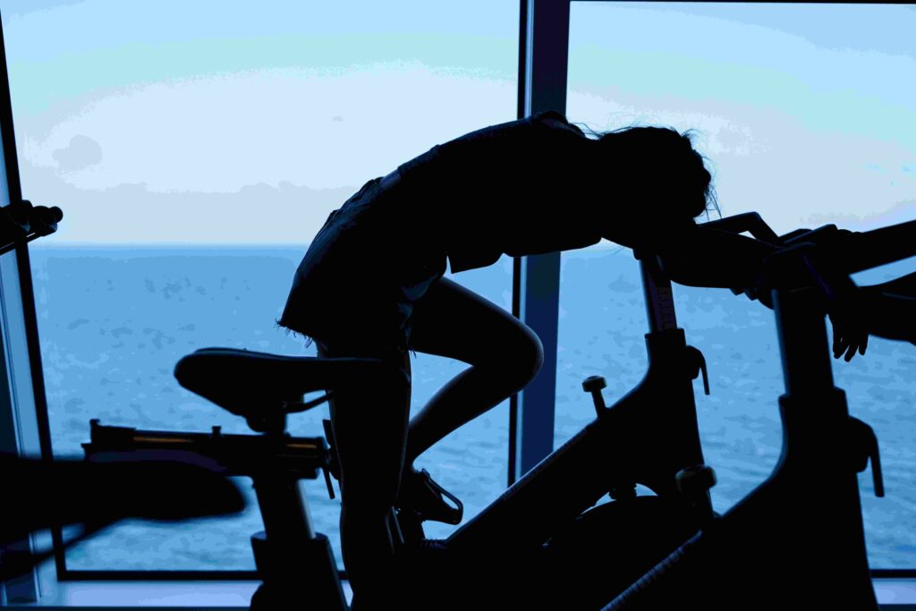 Pessoa exausta apoiada sobre uma bicicleta ergométrica em uma academia com vista para o mar, representando os efeitos do overtraining.