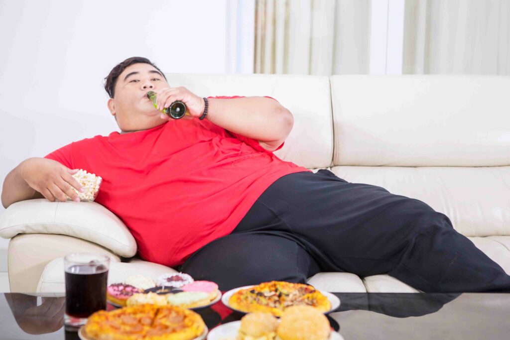 Homem obeso deitado no sofá comendo fast food e bebendo refrigerante, representando hábitos associados à Síndrome Metabólica.