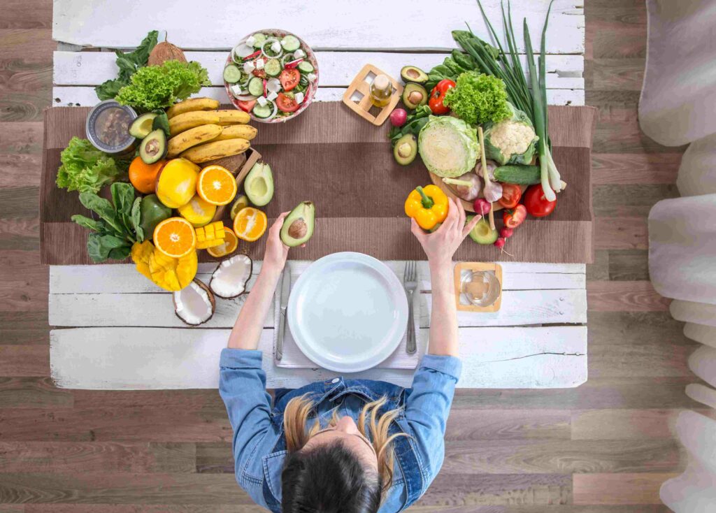 Pessoa vista de cima segurando um abacate e um pimentão amarelo diante de uma mesa com frutas, legumes e verduras frescas.