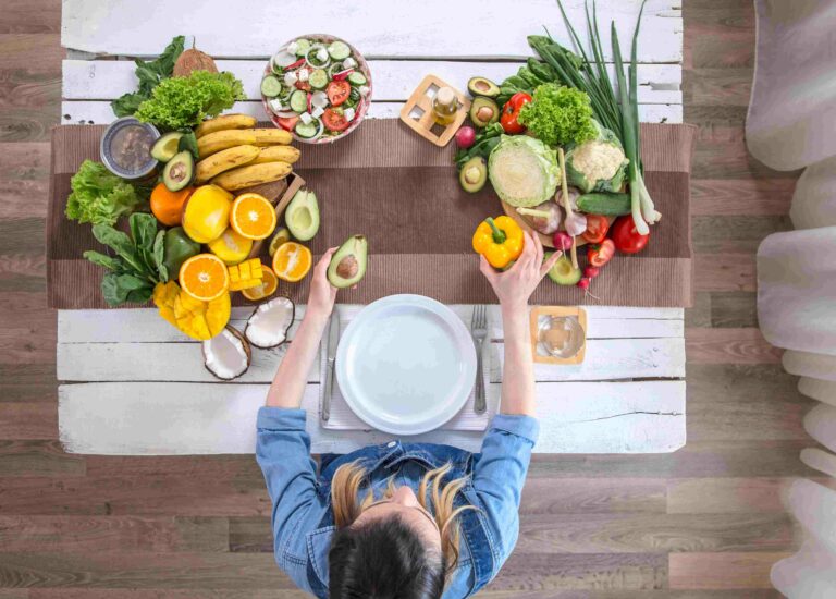 Pessoa vista de cima segurando um abacate e um pimentão amarelo diante de uma mesa com frutas, legumes e verduras frescas.