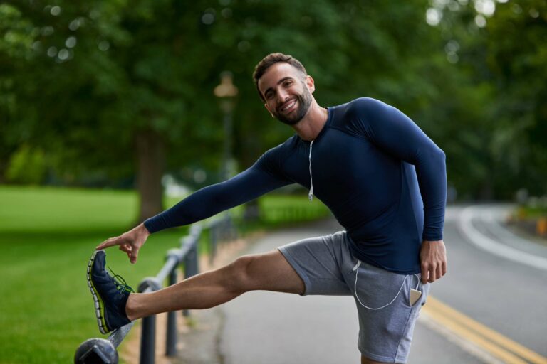 Homem fazendo alongamento na rua com roupa esportiva, ao lado de uma área arborizada.