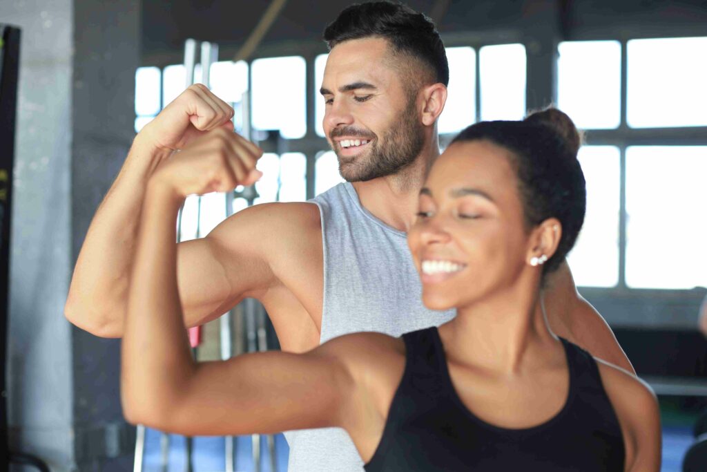 Homem e mulher sorrindo enquanto mostram os braços musculosos em uma academia, simbolizando saúde e força muscular.