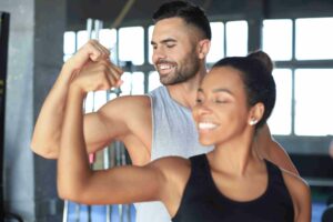 Homem e mulher sorrindo enquanto mostram os braços musculosos em uma academia, simbolizando saúde e força muscular.