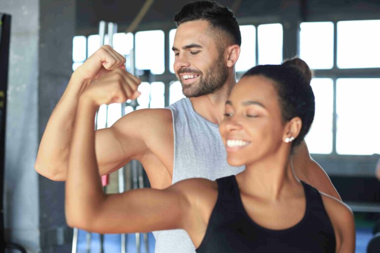 Homem e mulher sorrindo enquanto mostram os braços musculosos em uma academia, simbolizando saúde e força muscular.